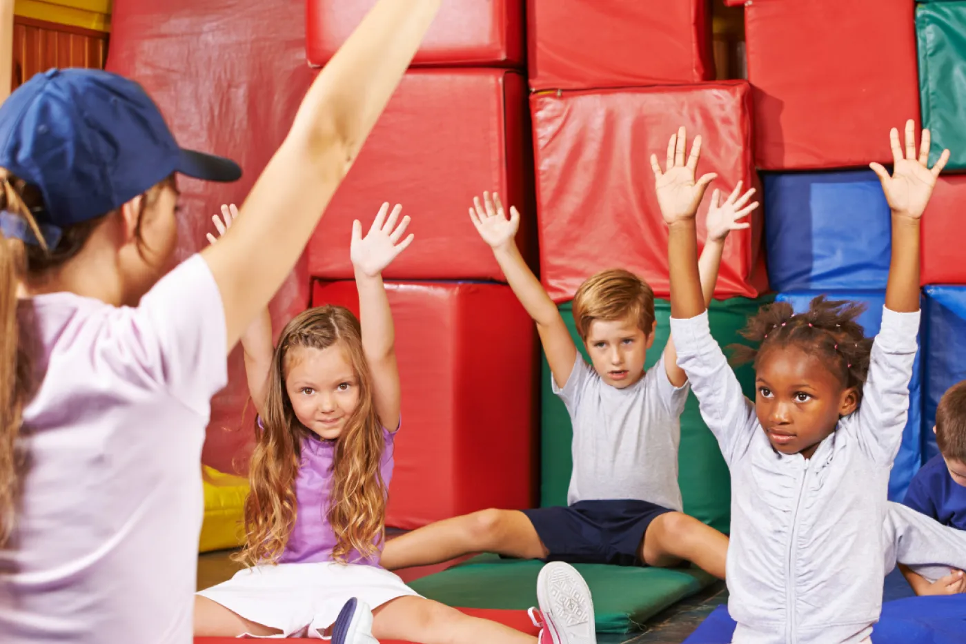 Kinder auf Turnmatten heben die Arme, angeleitet von einer Trainerin