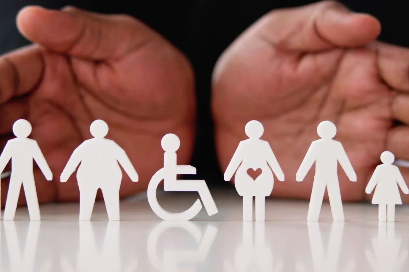 H&auml;nde liegen mit Handfl&auml;chen sichtbar auf einem Tisch, davor sind wei&szlig;e Figuren von Personen unterschiedlichsten K&ouml;rperbaus und im Rollstuhl aufgereiht.