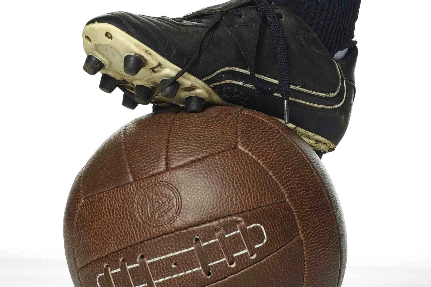 Fu&szlig; in schwarzem Fu&szlig;ballschuh steht auf altem braunem Leder-Fu&szlig;ball
