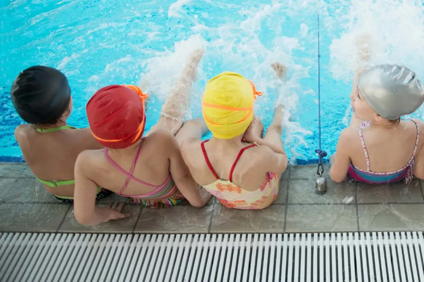 Vier Kinder in Badesachen und Schwimmkappen sitzen am Beckenrand. 