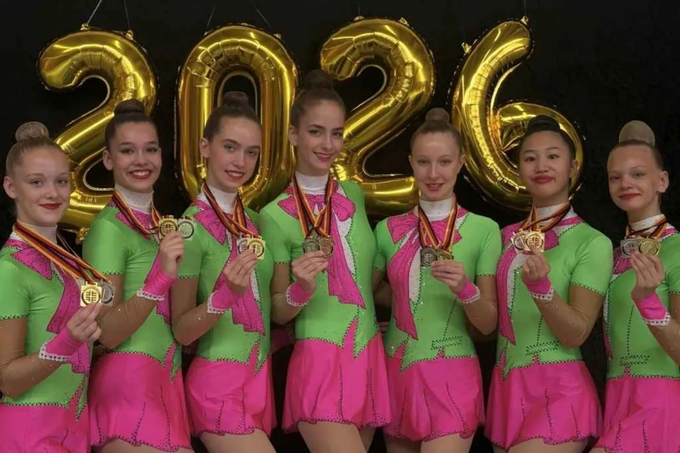 7 Frauen in pink-gr&uuml;nen Gymnastikanz&uuml;gen zeigen Medaillen im Kamera